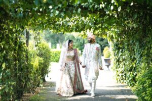A newlywed Indian couple in elegant wedding attire walks hand-in-hand under a leafy archway.