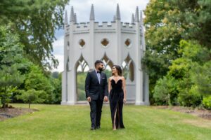 A stylish couple walks hand-in-hand in a grassy park with a white gothic structure behind them.