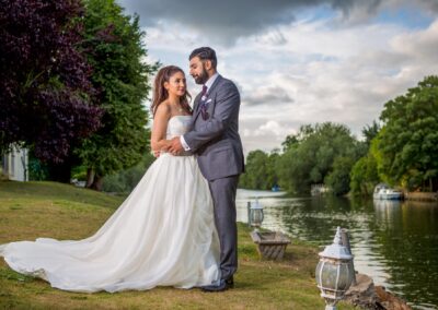 Bride and groom embracing beside tranquil river with trees and cloudy sky backdrop.