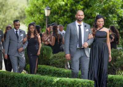 Wedding guests walking arm in arm in elegant black and grey outfits through a garden path.