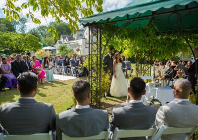 Outdoor wedding ceremony with couple under gazebo and guests seated in sunlight.