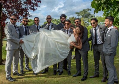 Groom and his groomsmen lifting the bride, smiling and dressed in suits, outdoors on a sunny day.