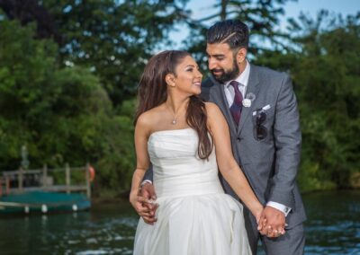 Bride and groom holding hands, standing close and smiling at each other in a garden by the river.