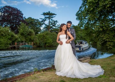 Bride and groom standing together by a river, with the groom holding the bride from behind as she smiles.