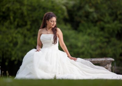 Bride in a strapless white gown sitting on a stone bench, looking down and smiling in a garden.