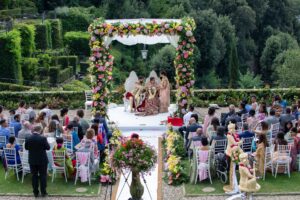 An outdoor wedding ceremony with a richly decorated arch and many guests.