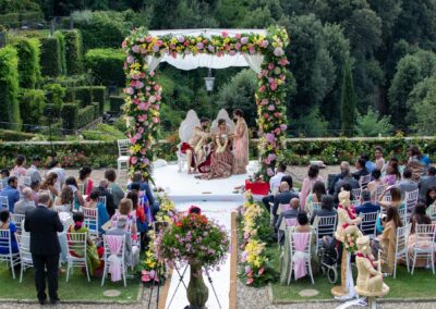 An outdoor wedding ceremony with a richly decorated arch and many guests.