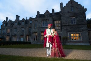 An Indian bride and groom stand outside a large, old stone manor house at dusk.