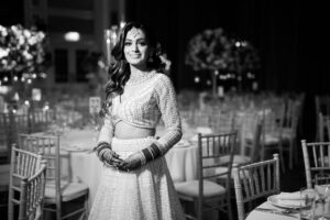 A black and white photo of a smiling Indian bride in a lehenga in a decorated hall.