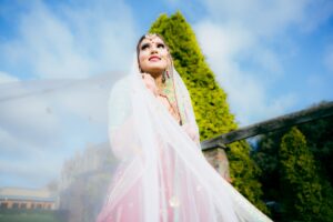A smiling Indian bride, veiled, looks upwards against a blue sky with green trees.