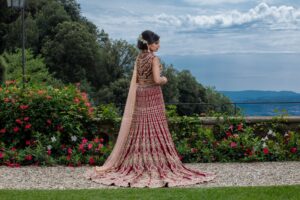 A bride in an elaborate red and gold gown stands outdoors, looking away.