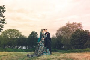 A newlywed couple in elaborate traditional attire stands embraced in a grassy field.