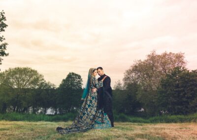 A newlywed couple in elaborate traditional attire stands embraced in a grassy field.