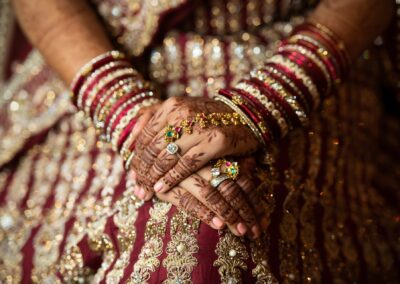 Close-up of bride’s mehndi hands with red bangles and gemstone rings on wedding dress.