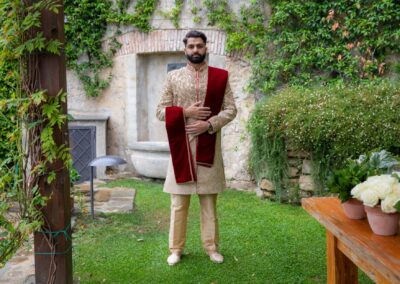 Groom standing in beige sherwani with red stole in a lush outdoor garden before wedding.