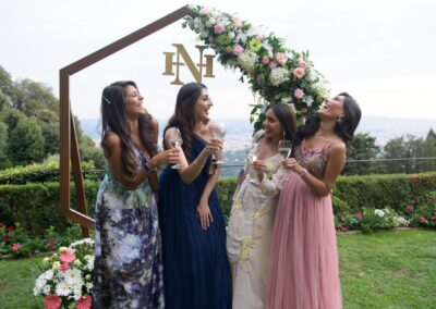 Four women holding glasses and laughing under floral arch at garden celebration.