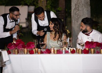 Two men sharing a laugh while handing microphone to bride during wedding toast.