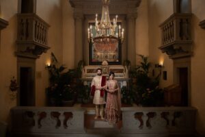 An Indian bride and groom pose formally under an ornate chandelier in a grand room.