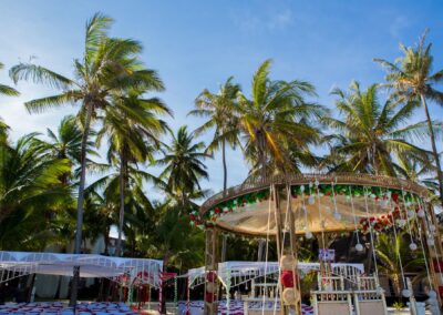 A decorated wedding canopy stands on a beach surrounded by tall palm trees.