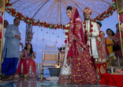 An Indian bride and groom stand smiling under a decorated canopy during their wedding ceremony.