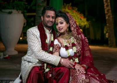 A newlywed Indian couple in elaborate traditional attire poses closely, smiling at the camera.