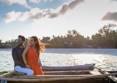 A couple relaxes on a traditional wooden boat on clear water with a tropical beach background.