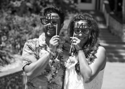 Couple wearing “Mr” and “Mrs” glasses posing playfully at tropical wedding party.