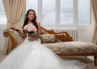 The bride, holding a bouquet, poses elegantly on a chaise lounge by a window.