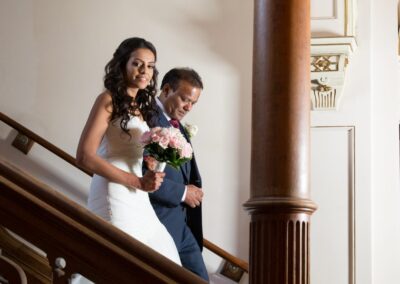 The bride, smiling, descends a grand wooden staircase with her father.