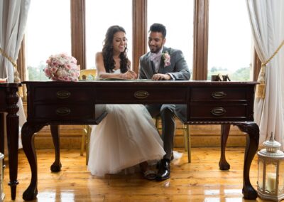 The bride and groom are seated at a wooden desk, signing documents by a bright window.