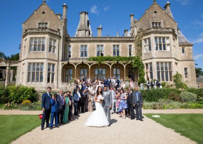 A large group of wedding guests and the couple pose in front of a grand manor.