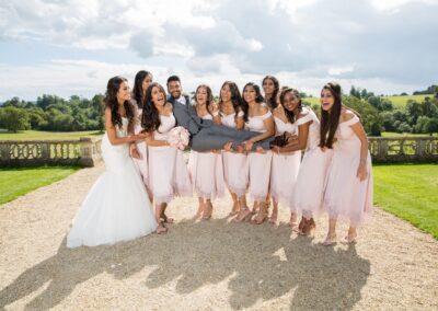 A playful wedding party, bridesmaids lifting the groom, all laughing outdoors.
