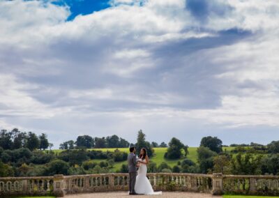 A couple holding champagne flutes, stands on a balcony overlooking a lush landscape.