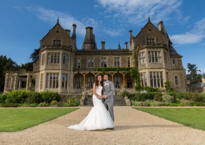 A newlywed couple, radiant, stands on a gravel path leading to a stately manor.