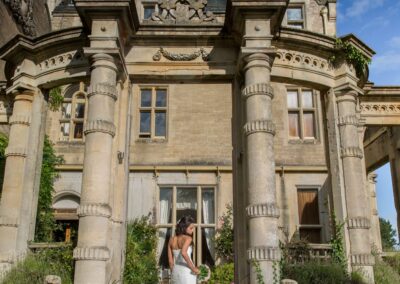A bride in a white gown stands elegantly on stone steps before a grand manor house.
