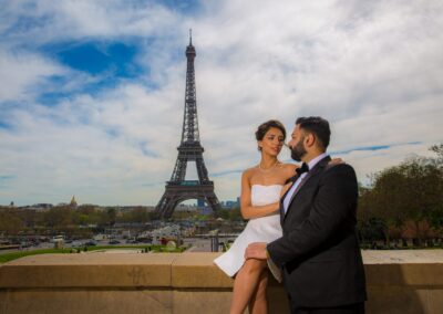 A couple embraces with the Eiffel Tower prominent in the background under a blue sky.
