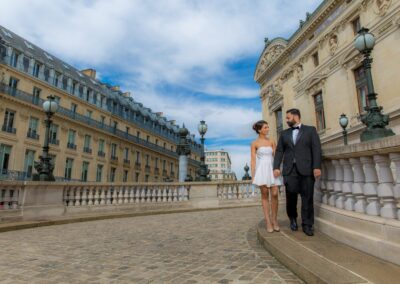 A couple in formal attire walks on a cobbled street in Paris with ornate buildings.