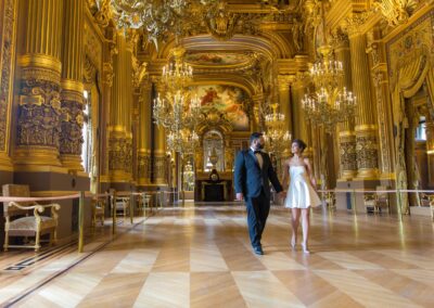 A couple walks hand-in-hand in a lavish, gold-adorned hall with chandeliers.