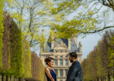 A romantic shot of a couple holding hands on a tree-lined path in front of a grand building.