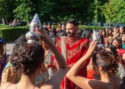 A groom in a red and gold sherwani is surrounded by women holding ceremonial items outdoors.
