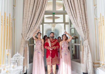 A groom in traditional Indian attire stands with two women in formal dresses at a doorway.