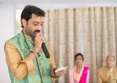 A man in traditional Indian attire speaks into a microphone at a wedding ceremony.