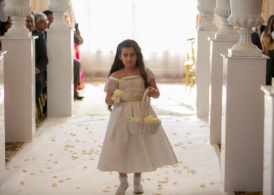 A young flower girl in a white dress holds a basket of petals and a flower as she walks the aisle.