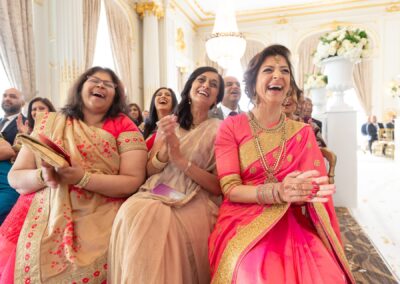 Three Indian women in traditional attire laugh and clap at a wedding.