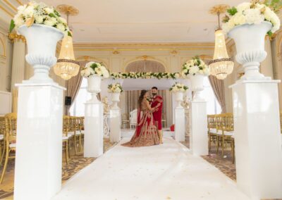 The bride and groom in traditional red outfits stand under a floral arch indoors.