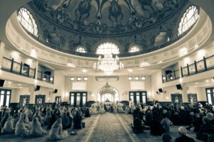 A wide shot of a large hall with people sitting on the floor, facing an altar and chandelier.