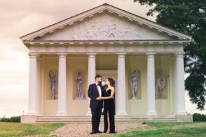 A couple in formal wear stands lovingly in front of a classical building with statues.