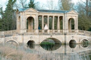 A couple stands on an ancient, arched stone bridge over water.