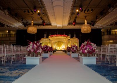 An empty wedding hall with a white aisle leading to an elaborate golden mandap and floral decor.