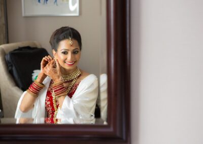 A bride in traditional attire looks in a mirror, adjusting her earring and smiling.